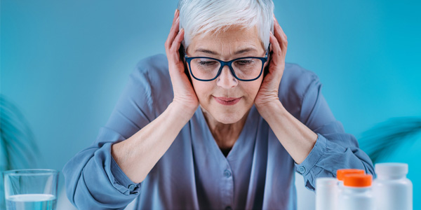 Eine Person sitzt an einem Tisch und hält mit beiden Händen den Kopf; auf dem Tisch stehen mehrere Medikamentenbehälter und ein Glas Wasser.